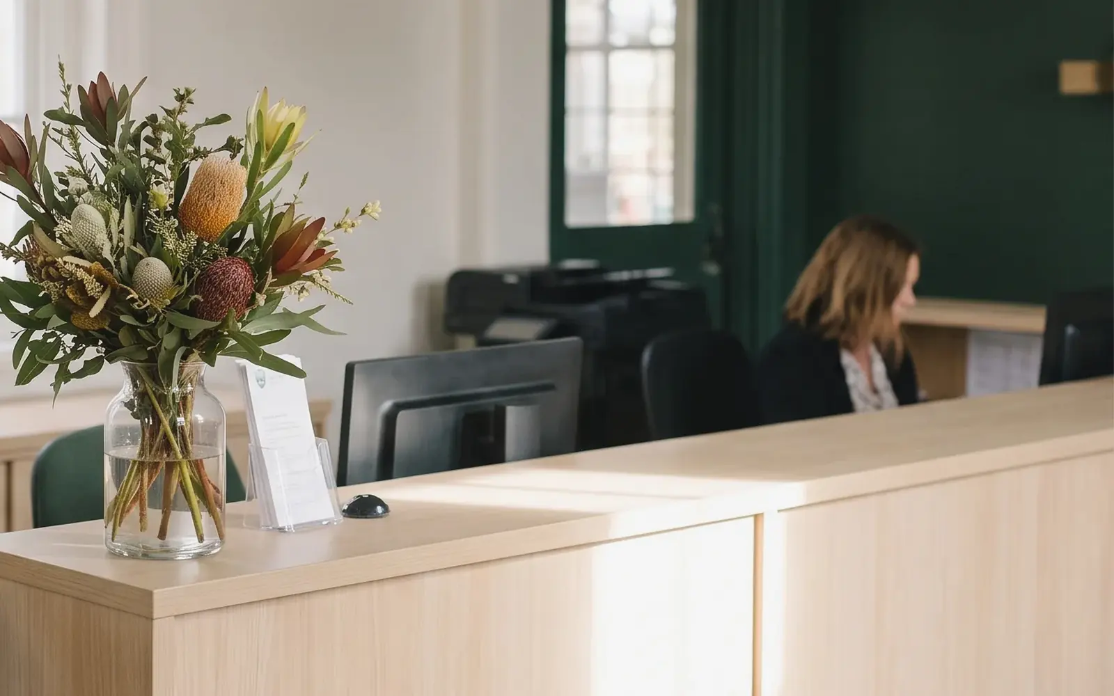 The front counter of an Australian school office, with a jug of native flowers, a brochure stand and a staff member working behind it.