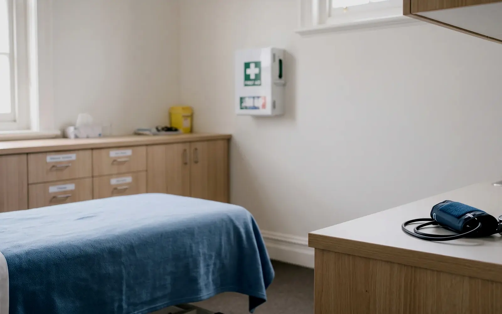 A school health centre with a made bed, a labelled cabinet of supplies and a wall-mounted first aid station.