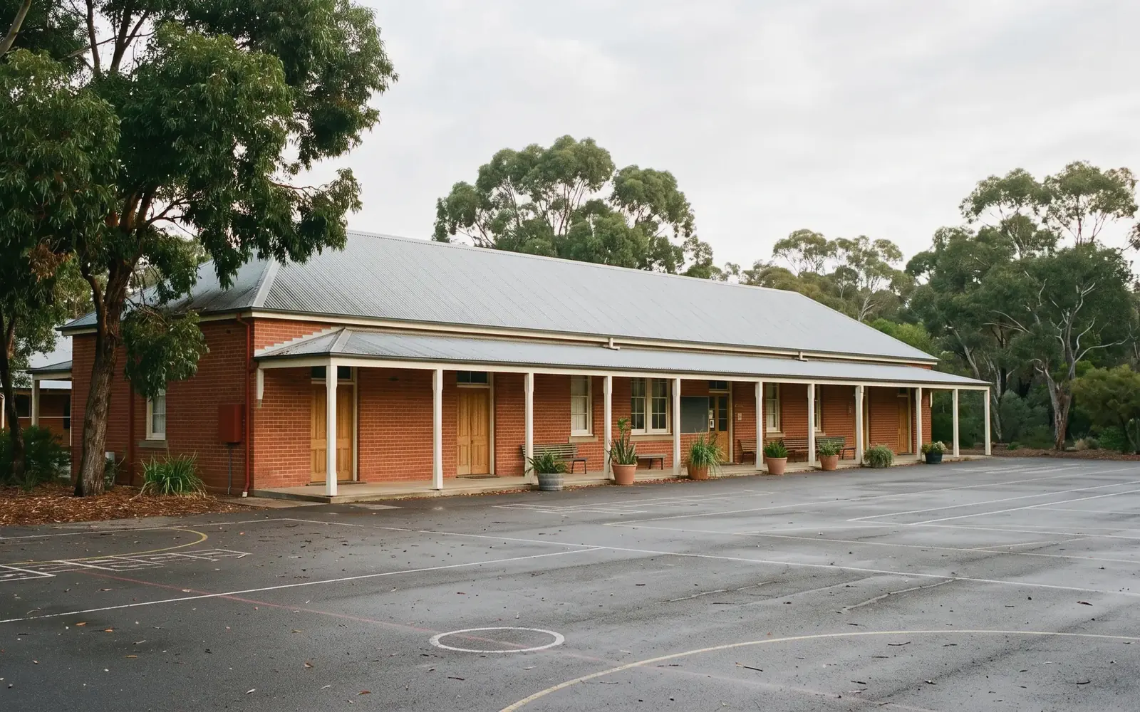 An established Australian school in red brick, with a verandah along its length, gum trees and an empty line-marked quadrangle.